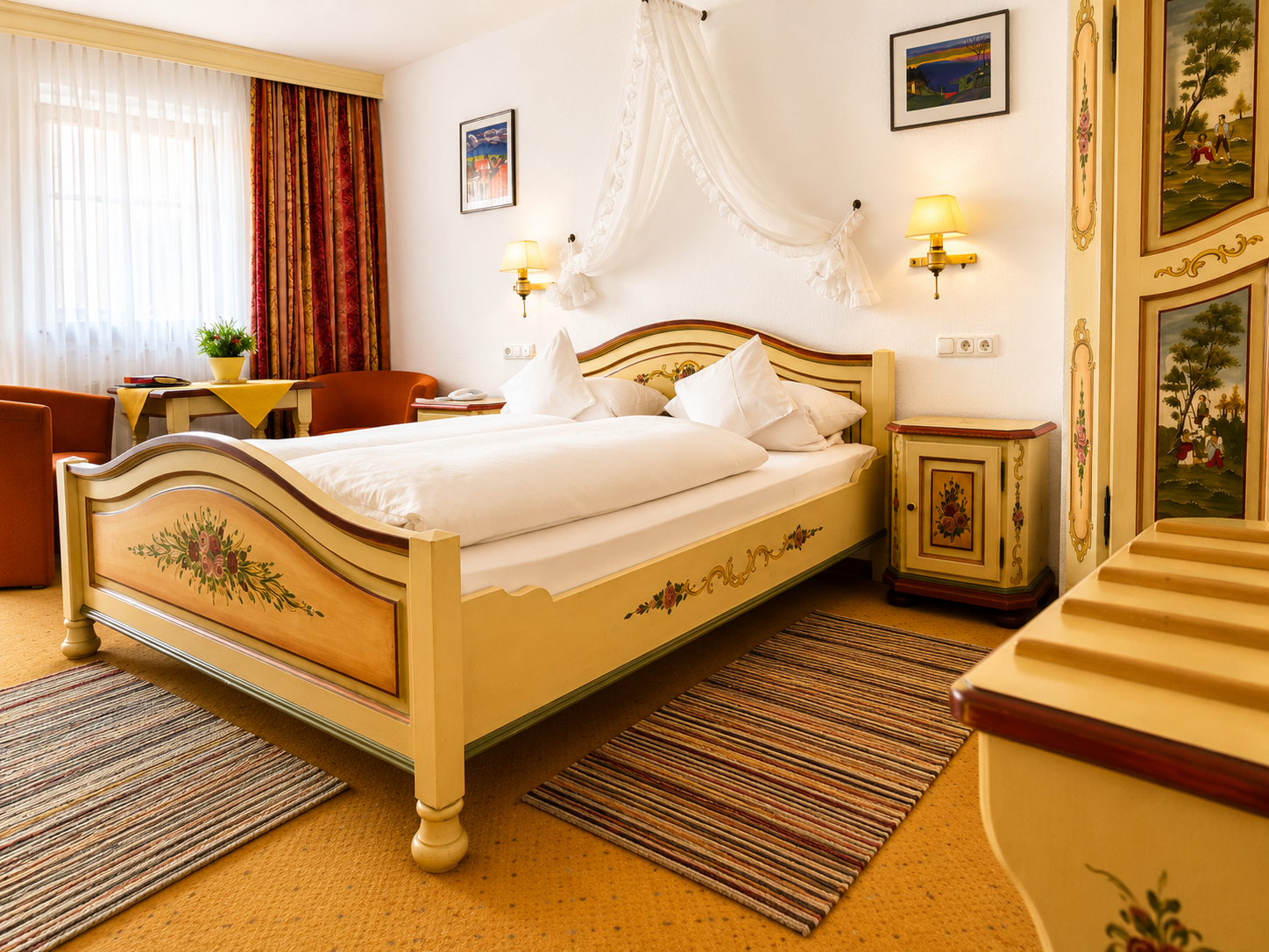 Comfort Doppelzimmer mit bemaltem Doppelbett, weißem Betthimmel, Nachttisch, Wandleuchten und Sesseln am Fenster