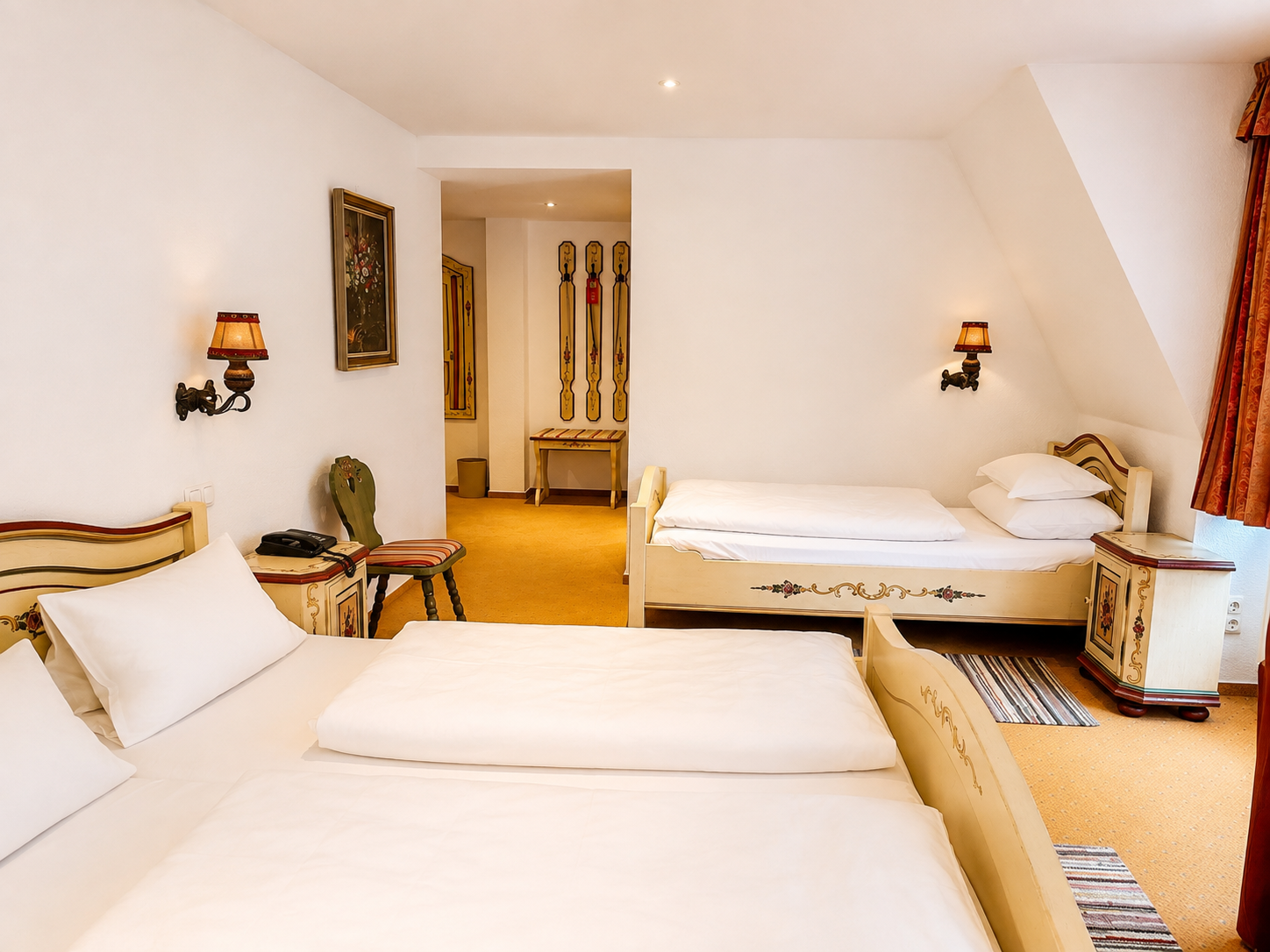 Dreibettzimmer mit Doppelbett im Vordergrund, einem bemalten Einzelbett dahinter, Nachttischen und Wandleuchten unter der Dachschräge