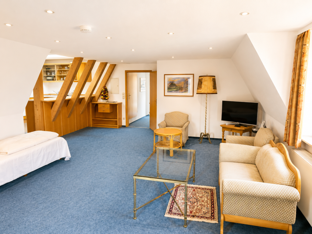 Living area under roof beams with a sofa, glass table, TV and single bed, with the kitchenette behind