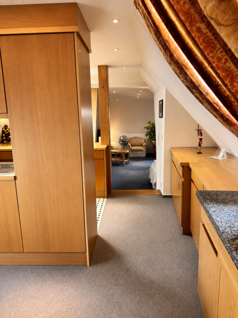 View through the kitchen area with light wooden cupboards into the living room with an armchair and a fan