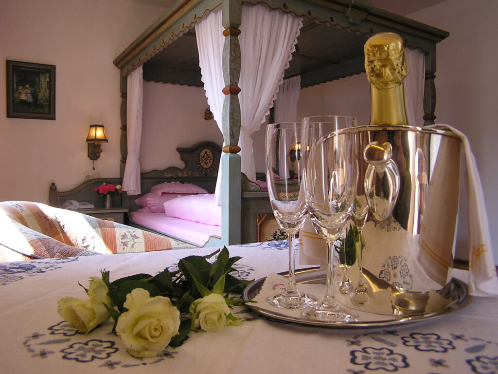 Sparkling wine cooler with a bottle and three glasses, and white roses on the bed, with the four-poster bed behind