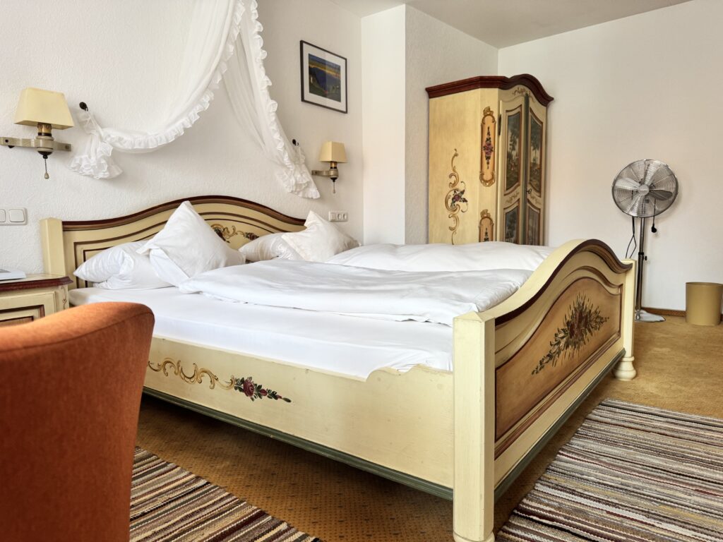Painted double bed with white bed linen, a painted farmhouse wardrobe, a standing fan and an orange armchair in the foreground
