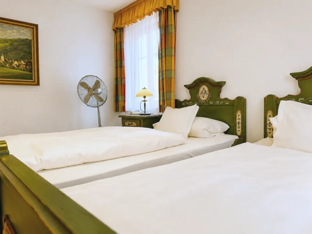 Zwei Einzelbetten mit weißer Bettwäsche, Nachttisch mit Lampe, Standventilator und Landschaftsbild an der Wand