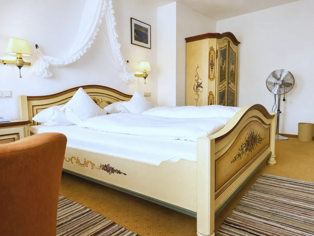 Painted double bed with floral decoration, behind it a painted farmhouse wardrobe, a standing fan and an orange armchair
